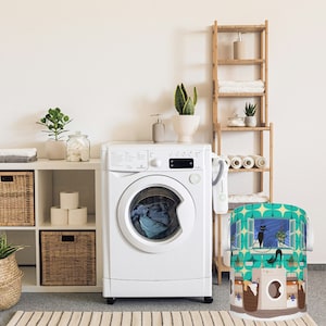 Könnte beinhalten: Eine weiße Waschmaschine mit einer Frontlader-Tür, ein Weiden-Wäschekorb mit einem bunten Design mit Katzen und Fenstern und ein Holzregal mit Handtüchern und Pflanzen.