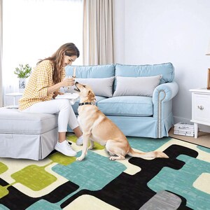 May include: A living room with a light blue sofa and a patterned rug with green, blue, black, and white shapes. A person sits on a gray ottoman, interacting with a dog. The room has a window with curtains and a side table.
