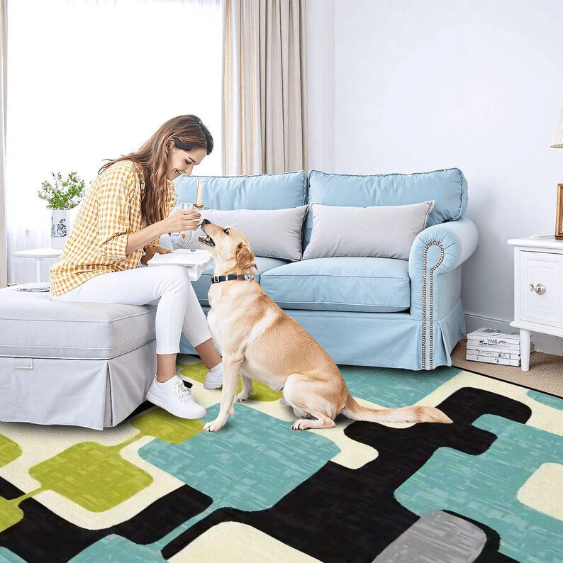 May include: A living room with a light blue sofa and a patterned rug with green, blue, black, and white shapes. A person sits on a gray ottoman, interacting with a dog. The room has a window with curtains and a side table.