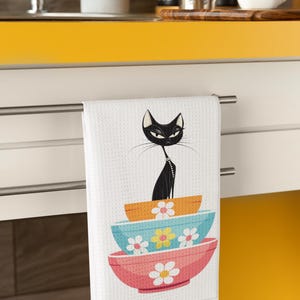 Puede incluir: Una toalla de cocina blanca con un gato negro sentado encima de tres cuencos coloridos con diseños florales. Los cuencos están apilados uno encima del otro y el gato mira hacia la derecha.