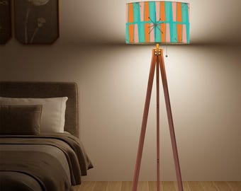 May include: A mid-century modern floor lamp with a wooden tripod base. The lampshade features a colorful pattern of vertical stripes in teal, orange, and pink, with a starburst design. The lamp is lit, casting a warm glow.
