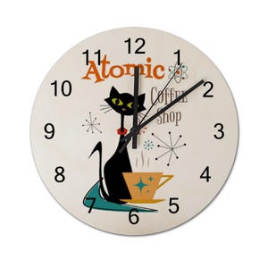 May include: A round, white clock with black hands and black numbers. The clock has a black cat with green eyes and a red collar sitting on a teal and blue shape. The cat is holding a yellow cup with a brown handle and a gold star. The clock also has the text "Atomic Coffee Shop" and a stylized atom symbol.
