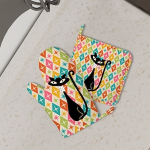 May include: A set of two oven mitts with a retro design featuring a black cat with a blue bow and a white pearl necklace. The mitts have a colorful geometric pattern with pink, orange, yellow, green, and blue stars and diamonds on a cream background.