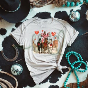 May include: A light beige t-shirt featuring a graphic of three horses with red bandanas, a pink bow, and heart-shaped balloons. The word "Cowgirl" is written in a cursive font. The shirt is styled with a knot at the waist.