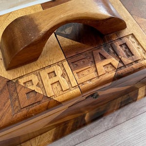 May include: A rectangular wooden bread box featuring a geometric parquet pattern of light and dark wood tones. The lid includes a curved wooden handle and the word BREAD carved into the surface in block capital letters across the top panel.