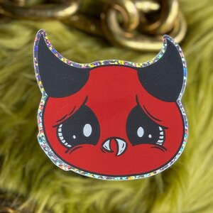 May include: A red and black glitter sticker of a cartoon devil with a sad expression. The devil has horns, a small mouth with a fang, and large eyes with tears.
