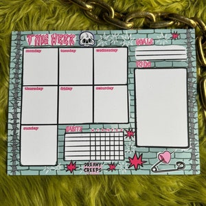 May include: A weekly planner with a green brick background and pink accents. The planner includes sections for goals, to-do lists, and daily tasks. It also features a habit tracker and a section for notes. The planner is decorated with a skull, stars, and a safety pin with a heart.