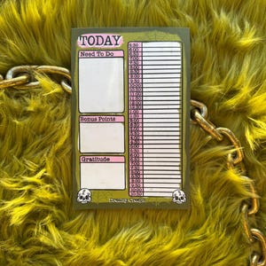 May include: A pink and green daily planner with the words "TODAY", "Need To Do", "Bonus Points", and "Gratitude" printed on it. The planner has a skull graphic and the text "Dreamy Creeps" at the bottom.
