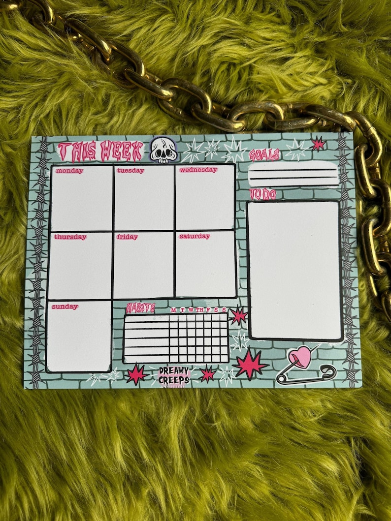 May include: A weekly planner with a green brick background and pink accents. The planner includes sections for goals, to-do lists, and daily tasks. It also features a habit tracker and a section for notes. The planner is decorated with a skull, stars, and a safety pin with a heart.