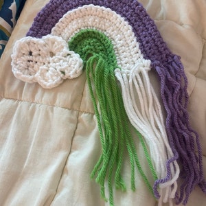 May include: A crocheted rainbow with purple, white, green, and white yarn. The rainbow has fringe on the bottom and a white crocheted cloud on the left side.