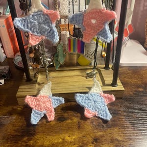 Puede incluir: Cuatro pendientes de estrella de ganchillo hechos a mano en blanco, azul claro y rosa. Los pendientes se exhiben en un expositor de joyas negro con otros collares y accesorios en el fondo. Las estrellas son una mezcla de colores.