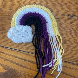 May include: A crocheted rainbow with a cloud at the top. The rainbow is made of yellow, white, purple, black, and white yarn. The yarn is tied in a fringe at the bottom.