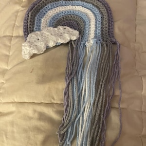 May include: A handmade crochet rainbow with a white cloud. The rainbow features shades of gray, blue, and white. The bottom of the rainbow has long, dangling yarn strands in coordinating colors. This item is perfect for nursery decor.