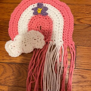 May include: A pink, white, and purple crocheted rainbow with a white crocheted cloud and pink and white yarn fringe.
