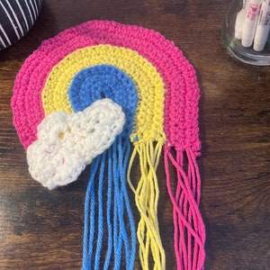 May include: A crocheted rainbow with a white cloud hanging from it. The rainbow is made of pink, yellow, blue, and white yarn. The yarn is tied into long strands that hang down from the bottom of the rainbow.