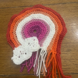 May include: A crocheted rainbow with a pink, white, and orange color scheme. The rainbow has a hole in the center and is surrounded by white crocheted flowers. The rainbow has long, colorful fringe.