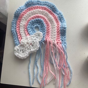 May include: A handmade crochet rainbow in light blue, pink, and white, with a white cloud and dangling yarn strands. The rainbow is a symbol of pride and support. The craft is made with soft yarn.