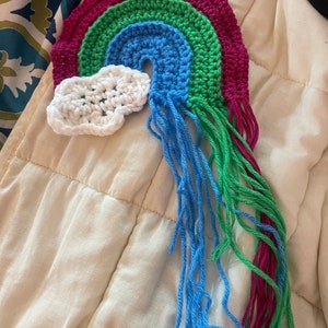 May include: A handmade crochet rainbow with a white cloud. The rainbow features bands of magenta, green, and blue, with blue and green fringe. The cloud is white. This item is likely a decorative piece or an accessory.