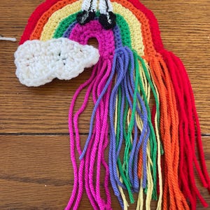 May include: A crocheted rainbow with a white cloud and rainbow colored yarn tassels. The rainbow is made of red, orange, yellow, green, blue, indigo, and violet yarn.