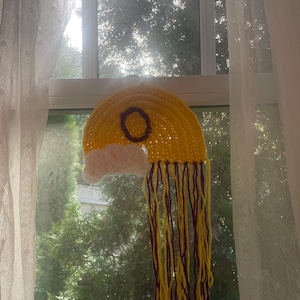 May include: A handmade, yellow crochet rainbow with a purple circle and white cloud accents. The rainbow has long yellow and purple fringe, hanging in front of a window with a view of trees and a sheer curtain.