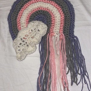 May include: A crocheted rainbow wall hanging with a cloud and tassels in shades of gray, pink, and white.