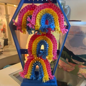 May include: Three crocheted rainbow ornaments featuring concentric arches of pink, yellow, and blue yarn with fringed ends. The ornaments are suspended from a blue, hexagonal-shaped wooden display stand with horizontal bars for hanging items.