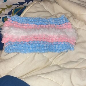 May include: A handmade headband featuring the colors of the transgender flag: light blue, pink, and white. The headband is made of a soft, textured material and has a fluffy white section in the middle.