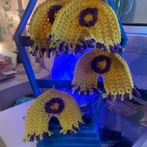 May include: Three yellow crocheted rainbow-shaped ornaments with purple circular centers and purple fringe along the bottom edges. The items are suspended from a blue hexagonal display stand with horizontal bars and small holes for hanging.