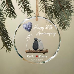 May include: A clear, round ornament with the text "Happy 20th Anniversary." It features a blue and white heart, and a couple made of stones sitting on a branch. The names "Emily" and "Kilyan" are below, along with the date 12.12.2005.