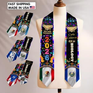 May include: Black graduation sash with colorful floral and butterfly designs, the text "2026", and flags of Mexico and El Salvador. The sash also includes the text "New York University" and "Carla Calderon B.S. in Biology".