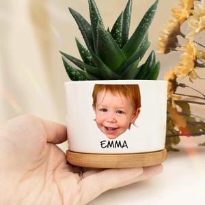 May include: White ceramic planter with a photo of a smiling child and the name "EMMA" printed on the front. The planter has a bamboo base and is holding a green succulent plant.