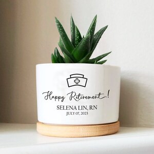 May include: White ceramic planter with a wood base. The planter has a black nurse's cap icon and the text "Happy Retirement! SELENA LIN, RN JULY 07, 2023". A green succulent plant is in the planter.