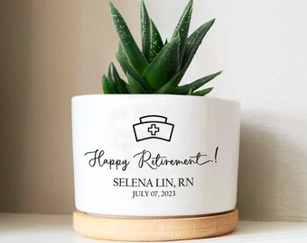 May include: White ceramic planter with a wood base. The planter has a black nurse's cap icon and the text "Happy Retirement! SELENA LIN, RN JULY 07, 2023". A green succulent plant is in the planter.