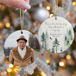 May include: A white ceramic ornament with a watercolor forest scene and the text "Those we love don't go away, they walk beside us every day". The ornament also includes the name "William James" and the dates "1952-2024". A second ornament features a photo of a man wearing a brown jacket and a hat.