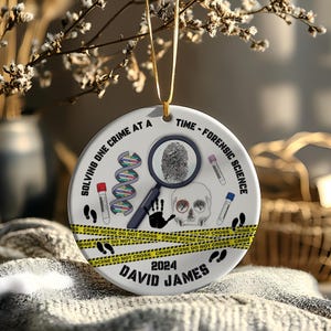 May include: A white ceramic ornament with a forensic science theme. It features a fingerprint, DNA strands, a skull, and the text "SOLVING ONE CRIME AT A TIME - FORENSIC SCIENCE". The ornament includes the name "DAVID JAMES" and the year "2024".