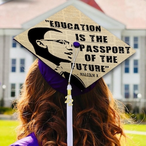 May include: A purple graduation cap featuring a beige background with a newspaper print pattern. It displays a black silhouette of Malcolm X and the quote, "EDUCATION IS THE PASSPORT OF THE FUTURE" - MALCOLM X. A white tassel with a gold "22" charm is attached.
