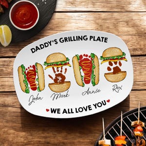 May include: White ceramic serving platter with the text "Daddy's Grilling Plate" and "We All Love You". The platter features four food items: a hot dog, a hamburger, a hot dog, and a hamburger. Each food item has a handprint on it and a name underneath: John, Mark, Annie, and Rex.