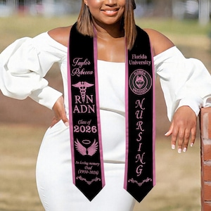 May include: A black graduation sash with pink text and accents. The sash features the text "Taylor Rebecca", "RN ADN", "Class of 2026", and "In loving memory Dad (1950-2026)". The other side says "Florida University" and "NURSING".