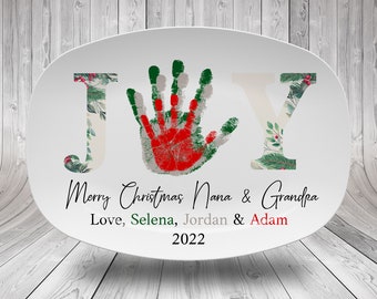 May include: White oval ceramic platter with a green, red and white handprint in the centre. The word "JOY" is written in green and white with floral accents. The text "Merry Christmas Nana & Grandpa Love, Selena, Jordan & Adam 2022" is written below the handprint.