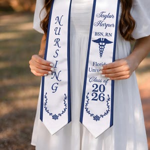 May include: White graduation sash with navy blue trim and text. One sash reads "NURSING" in navy blue. The other sash reads "Taylor Harper BSN, RN" with a caduceus symbol, "Florida Univ" and "Class of 2026".