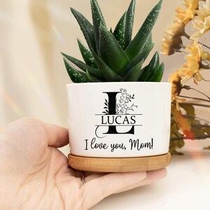 May include: White ceramic planter with a bamboo base. The planter has a floral design with the letter L and the name Lucas. The planter also has the text "I love you, Mom!"