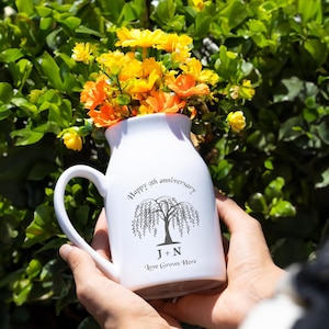 May include: White ceramic pitcher with a handle, filled with yellow and orange flowers. The pitcher has a black tree design and the text "Happy 9th anniversary" and "J + N Love Grows Here".
