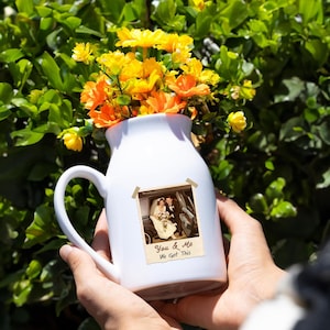 May include: A white ceramic pitcher filled with yellow and orange flowers. A vintage photograph is taped to the front of the pitcher, with the text "You & Me We Got This". The pitcher has a handle and is held in front of a green leafy background.