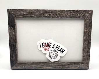May include: A framed print with a distressed, dark brown wooden frame. The print features a white sticker with the text "I HAVE A PLAN" and a die graphic. The word "HAD" is crossed out in red.