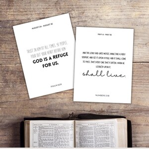 May include: Two white cards with Bible verses and dates, "August 24 - August 30" and "May 4 - May 10", sit on a wooden surface. The text on the cards includes "God is a refuge for us" and "shall live". An open Bible is at the bottom.