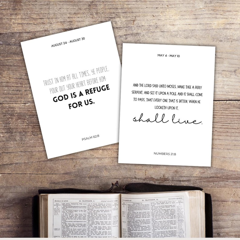 May include: Two white cards with Bible verses and dates, "August 24 - August 30" and "May 4 - May 10", sit on a wooden surface. The text on the cards includes "God is a refuge for us" and "shall live". An open Bible is at the bottom.