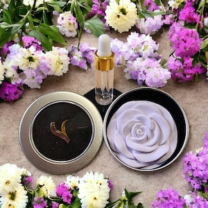 Può includere: Una scatola rotonda con un sapone a forma di rosa viola all'interno. La scatola è aperta e il sapone è visibile. Una piccola bottiglia di olio essenziale è sullo sfondo.