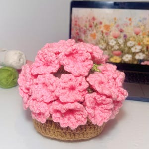 Puede incluir: Arreglo floral tejido a ganchillo en una cesta marrón. Las flores son de un rosa brillante, con pétalos detallados. La cesta tiene un aspecto texturizado y tejido. Un portátil con un fondo de pantalla floral y lana se encuentran en el fondo.