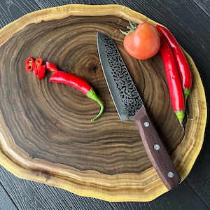 Puede incluir: Una tabla de cortar de madera con borde natural, con un cuchillo, chiles rojos y un tomate. El cuchillo tiene una hoja oscura estampada y un mango marrón. También hay chiles cortados en la tabla.