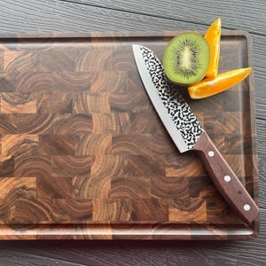 May include: A rectangular end-grain wooden cutting board with a dark brown, multi-tonal block pattern. A kitchen knife with a hammered metal blade and a dark wood handle rests on the board next to a sliced green kiwi and orange citrus wedges.